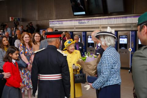 女王陛下身穿黄色外套，头戴黄色帽子, 和一个穿红毛衣的女学生, 穿蓝/红制服的人, 一位戴着帽子的蓝衣女士和其他公众.