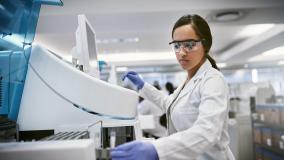Medium skinned woman with dark hair in a lab coat, safety glasses and blue gloves works in a lab environment
