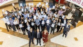 View from balcony looking down at the graduate cohort holding certificates in the air