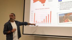Man in white t-shirt and check shirt pointing to charts on screen
