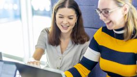 Brunette girl in light blue button-up shirt looks on a laptop with a blond girl in glasses and a navy and gold striped shirt