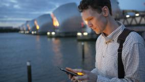 White man in a button up shirt looks at a cellphone with the Thames Barrier in the background