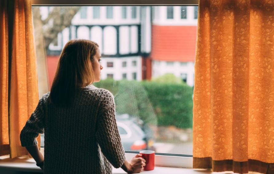 Stock image of woman in a beige sweater looking out a window draped with gold curtains