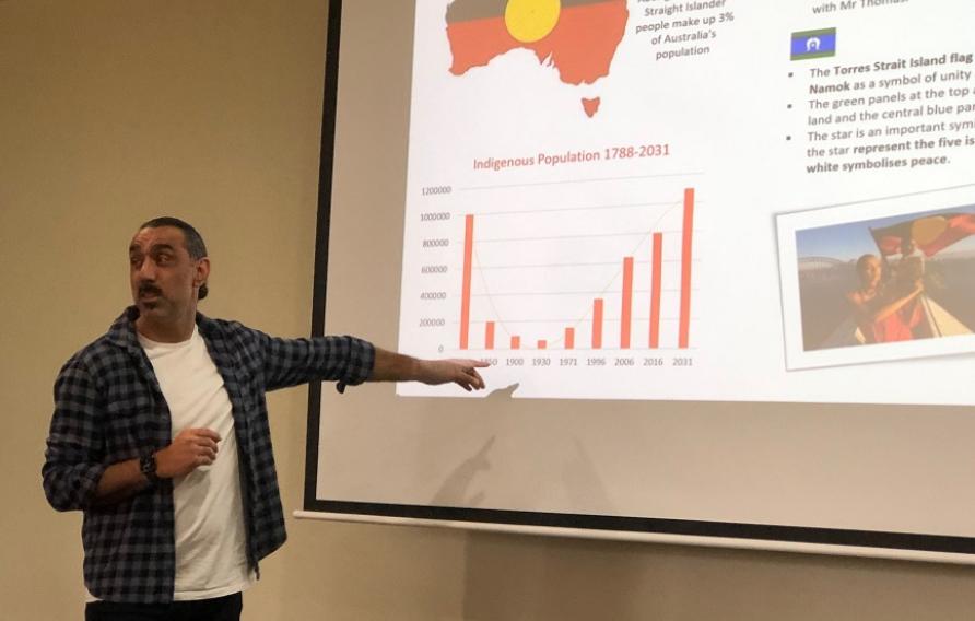 Man in white t-shirt and check shirt pointing to charts on screen