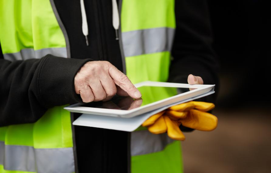 Man in yellow PPE vest holds yellow work gloves and uses an iPad