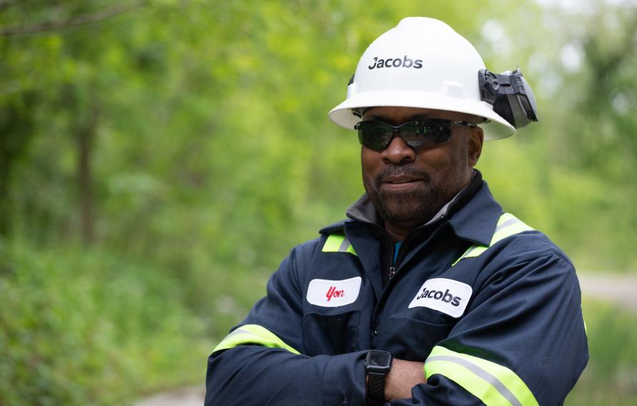 Black man in a navy PPE suit with a white Jacobs hardhat poses in front of greenery