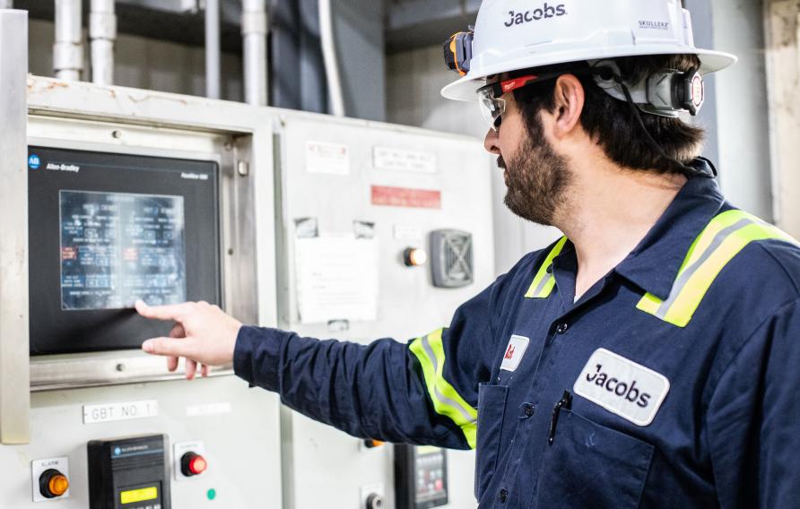 Dark haired man in navy jump suit and white hard hat points at a machine monitor