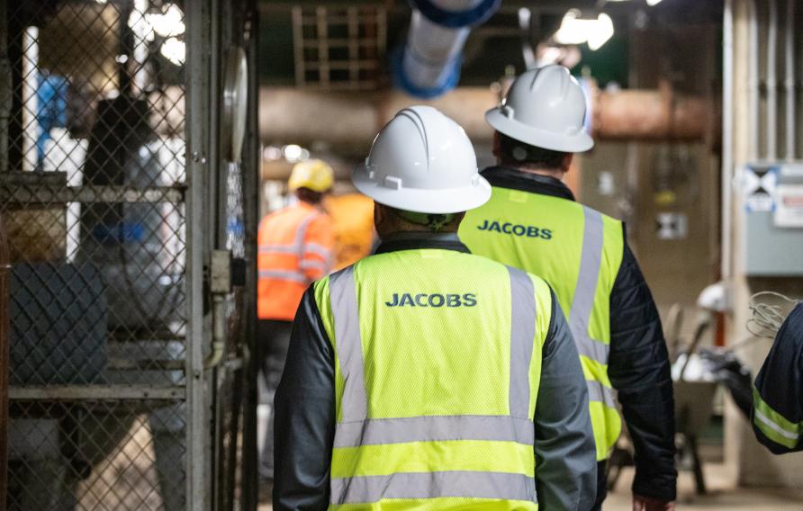 Two men in yellow Jacobs PPE vests and white hard hats walk into a control room