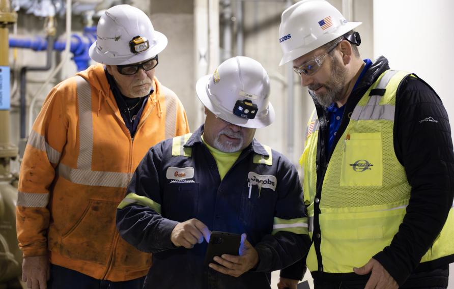 Three men at various heights in different colored PPE look on an iPad