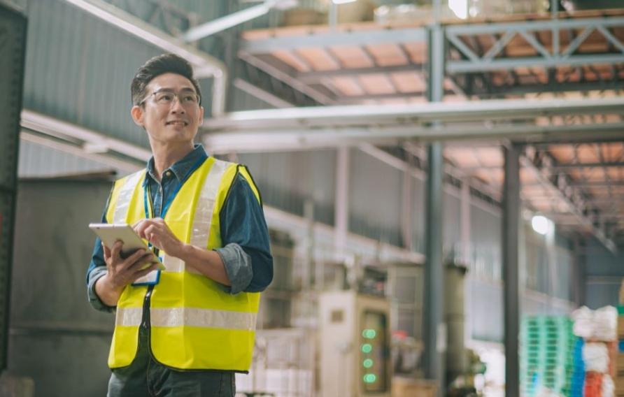 Person in a high-visibility vest holding a tablet in a large industrial warehouse