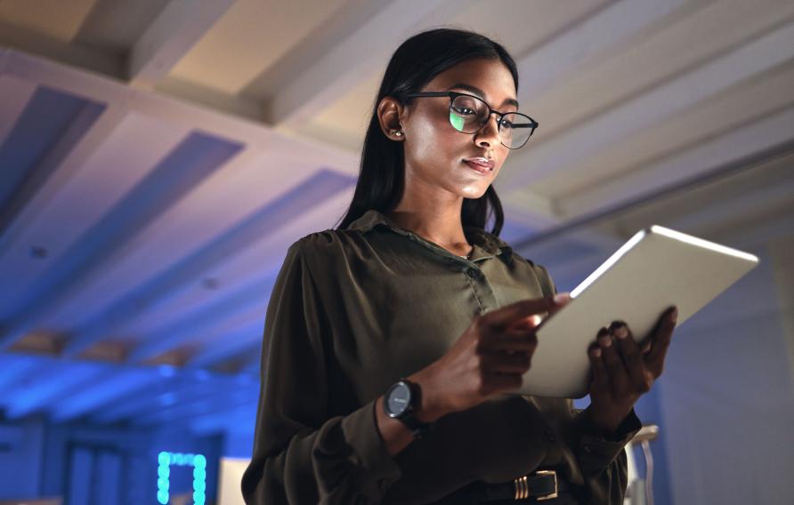 Tablet, designer and serious woman research in business startup office at night on deadline. Technology, creative professional or Indian entrepreneur reading information, email or app in neon company 