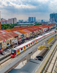 Elevated red and yellow rail cars with a cityscape in the background and red roofed apartment-style residences passing on the left