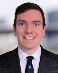 Man smiling with brown short hair, dressed in white shirt, blue tie and check jacket.