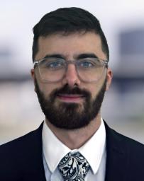 Man with dark brown hair, beard, wearing glasses, blue jacket and white shirt plus tie.