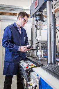 White man in safety glasses and blue lab coat works on a test rig in a lab