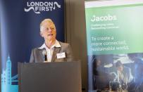 Woman speaking, stood behind lectern, with banners behind her.