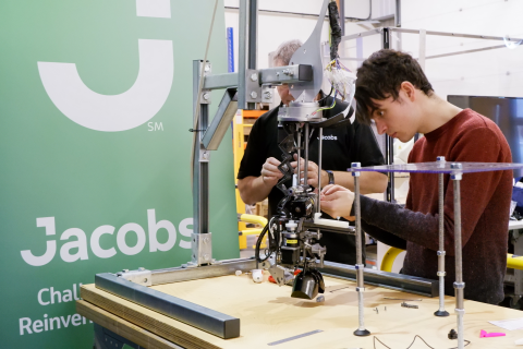 Dark haired man uses a robot in front of a green Jacobs banner