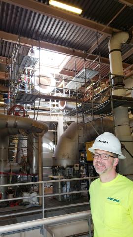 Man in yellow t-shirt with hard hat smiling in front of pipes