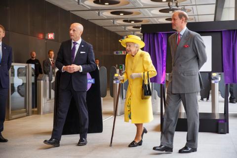 Her Majesty the Queen dressed in yellow coat and hat walking with TfL Commissioner Andy Byford on her right and Prince Edward, the Earl of Wessex on her left