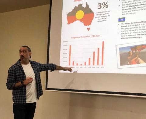 Man in white t-shirt and check shirt pointing to charts on screen