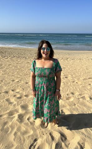Dark haired woman in sunglasses and a green dress poses on a beach with ocean in the background
