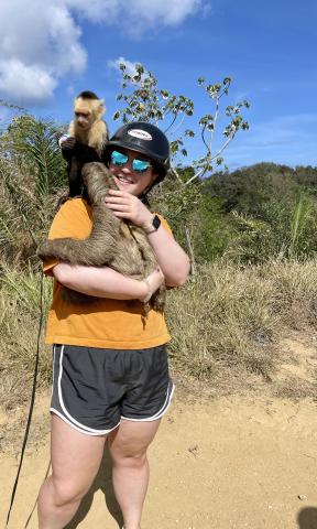 Woman in orange shirt and grey shorts with a hat and sunglasses holds a sloth 