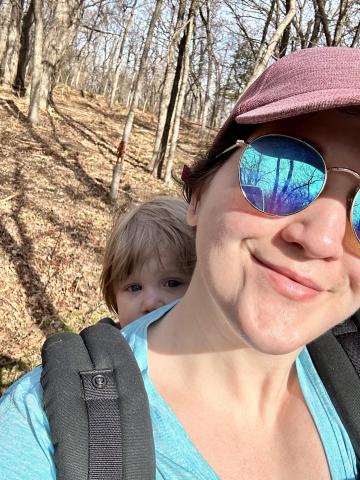 Woman in sunglasses and a red hat with a baby in a backpack carrier hikes in the woods