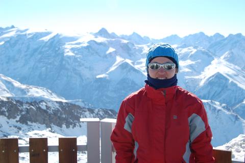 Woman in red ski jacket and blue cap poses in front of a snow-capped mountain