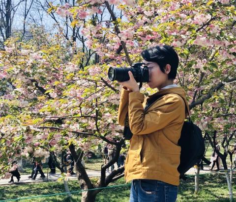 Woman with dark black hair in a yellow coat takes a photo with a black camera in front of blooming trees