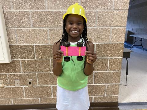 Young Black girl in a yellow plastic hard hat proudly smiles with her hot pink race car