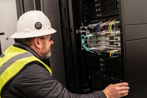 Bearded man in white hard hat and yellow PPE vest looks at a data system