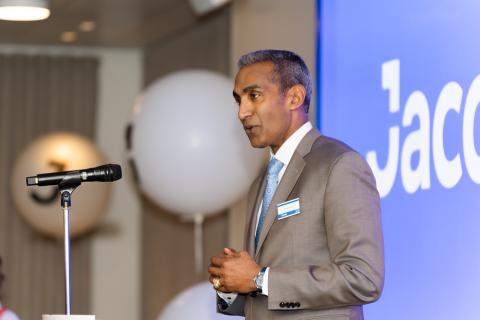 Man in beige suit, white shirt and blue tie speaking at microphone