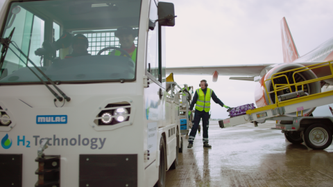 Man in blue trousers and hi vis vest helps to load luggage onto aircraft.