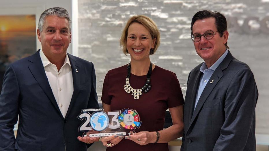 Two white men flanking a blond woman in a maroon dress and statement necklace holding a 2030 trophy