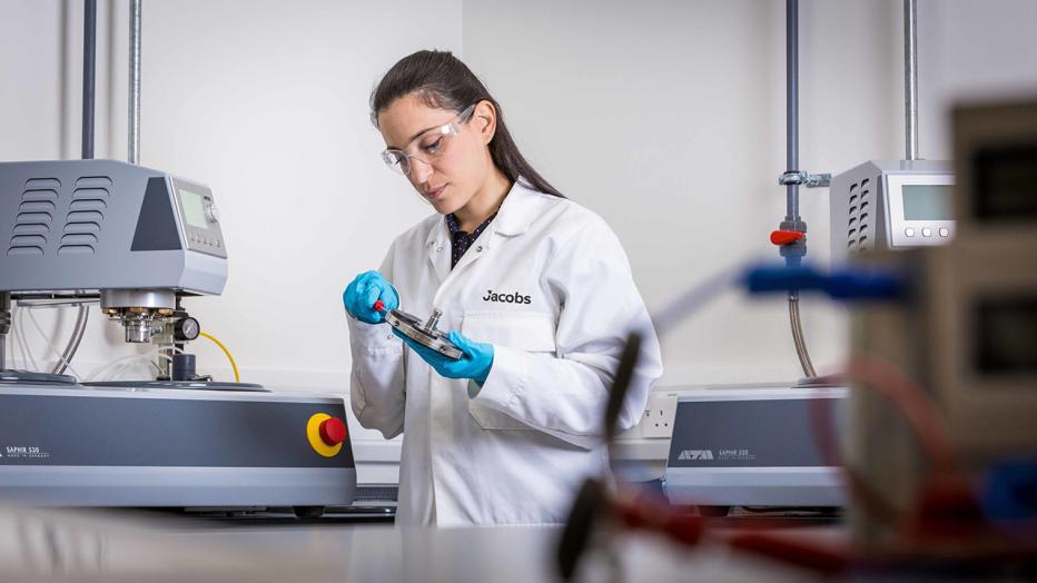 Woman in long black hair in a white lab coat and safety glasses in lab