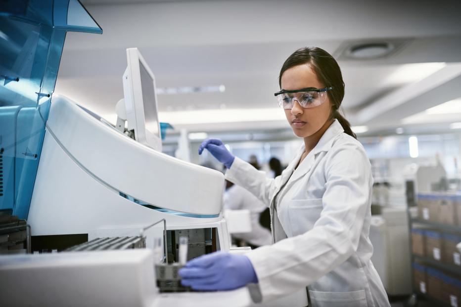 Medium skinned woman with dark hair in a lab coat, safety glasses and blue gloves works in a lab environment