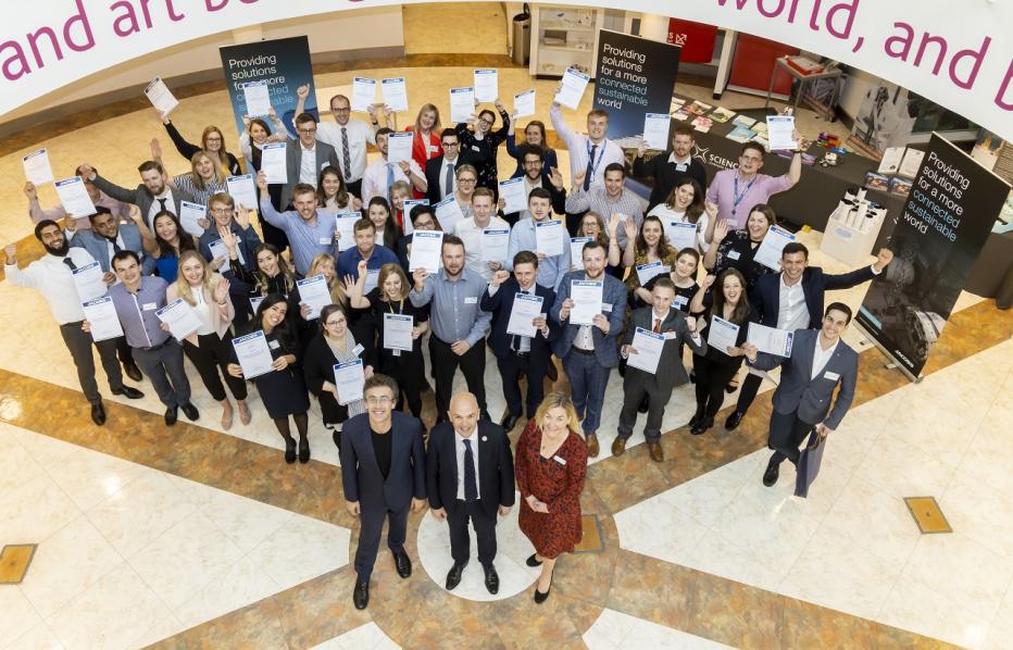 View from balcony looking down at the graduate cohort holding certificates in the air