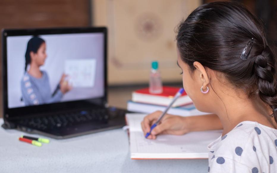 Girl writing in notebook looking at virtual lesson on laptop screen