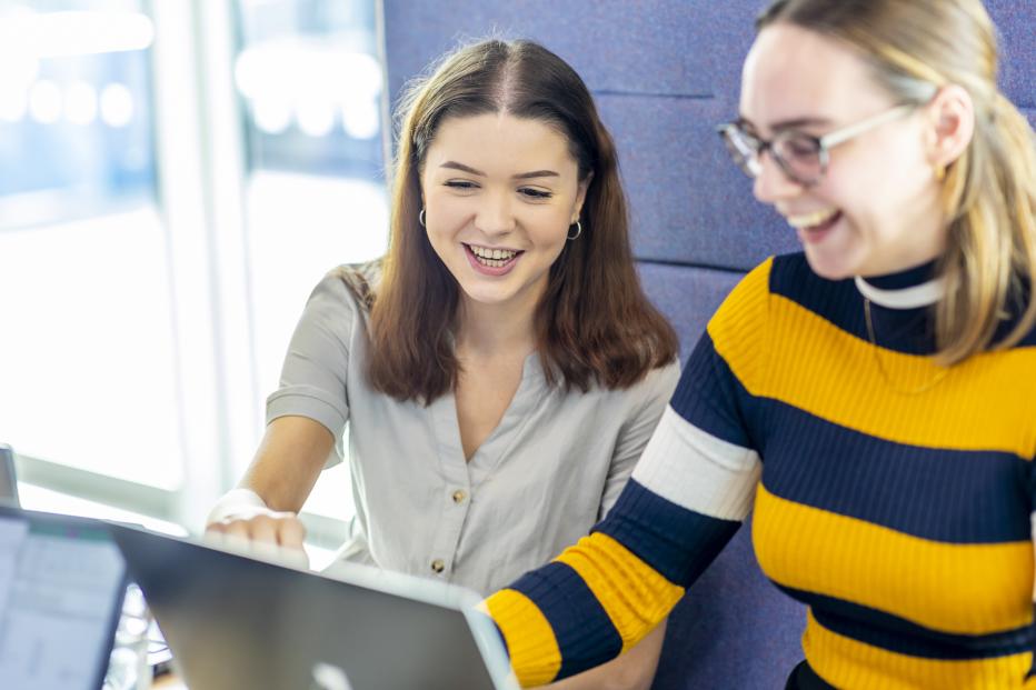 Brunette girl in light blue button-up shirt looks on a laptop with a blond girl in glasses and a navy and gold striped shirt