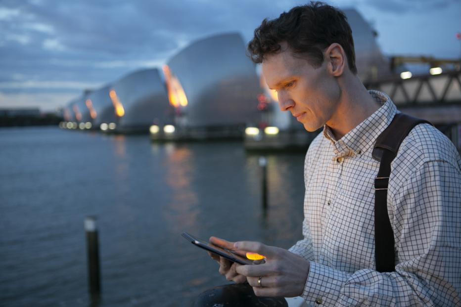 White man in a button up shirt looks at a cellphone with the Thames Barrier in the background