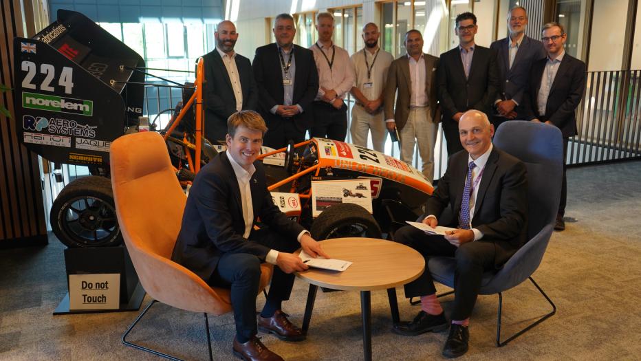Two men sat at round table with documents and group of men behind.