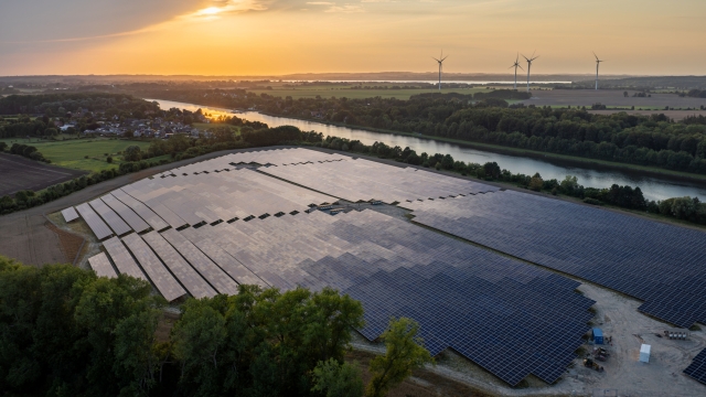 Aerial view of an extensive solar panel installation near a river, with wind turbines and farmland in the background at sunset.