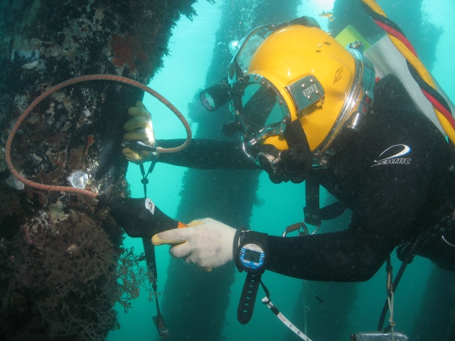 Underwater diver wearing a hard helmet and protective gloves examines a submerged structure with inspection tools.