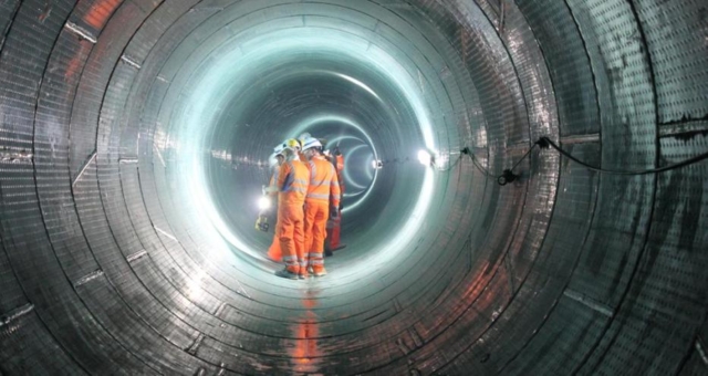 Several construction personnel wearing high‑visibility clothing stand inside a circular underground tunnel.