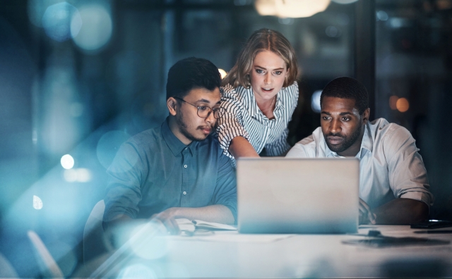 Three people gathered around a laptop at a table in a dimly lit office, reviewing information on the screen.