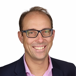 Studio‑style head‑and‑shoulders portrait of a person wearing rectangular glasses, a dark blazer, and a light pink collared shirt, photographed against a plain white background.