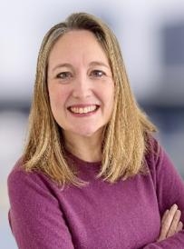 Professional upper‑body portrait of a person wearing a maroon crew‑neck top, photographed with arms crossed against a softly blurred indoor background.