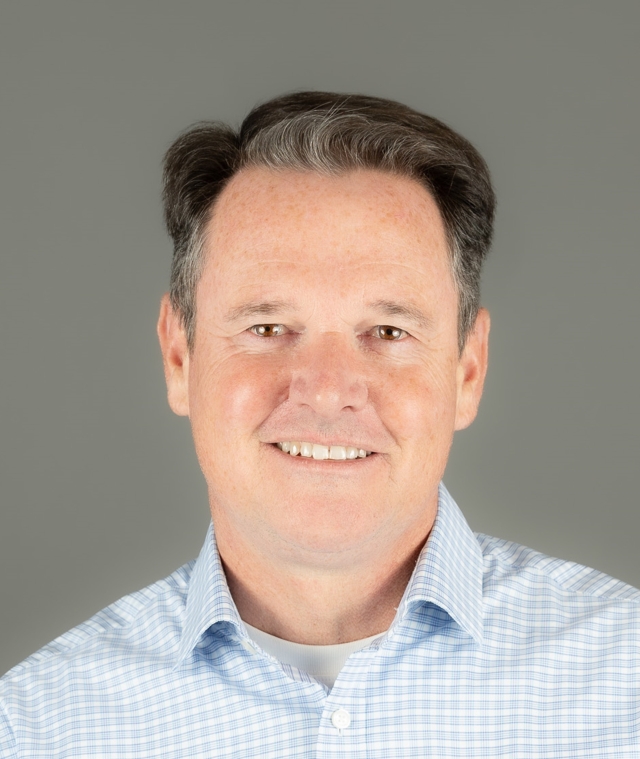 Studio‑style head‑and‑shoulders portrait of a person in a light blue checkered button‑down shirt, photographed against a plain gray background.