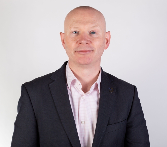 Head‑and‑shoulders studio portrait of a person in a dark business jacket over a light pink collared shirt, photographed against a plain white background.