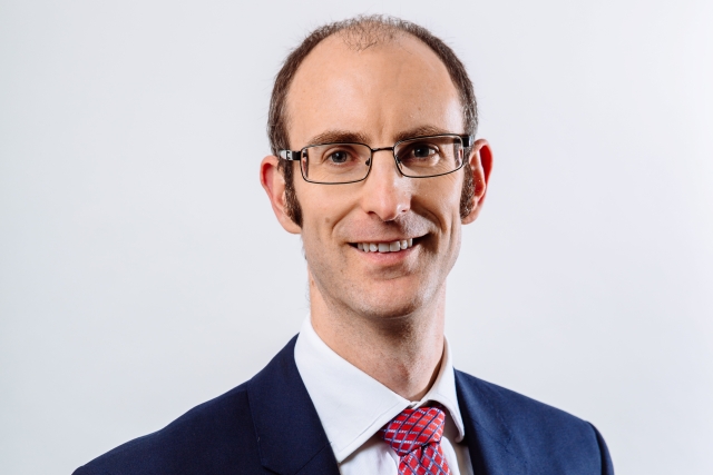 Studio‑style head‑and‑shoulders portrait of a person wearing rectangular glasses, a dark suit jacket, white collared shirt, and red patterned tie, photographed against a light gray background.